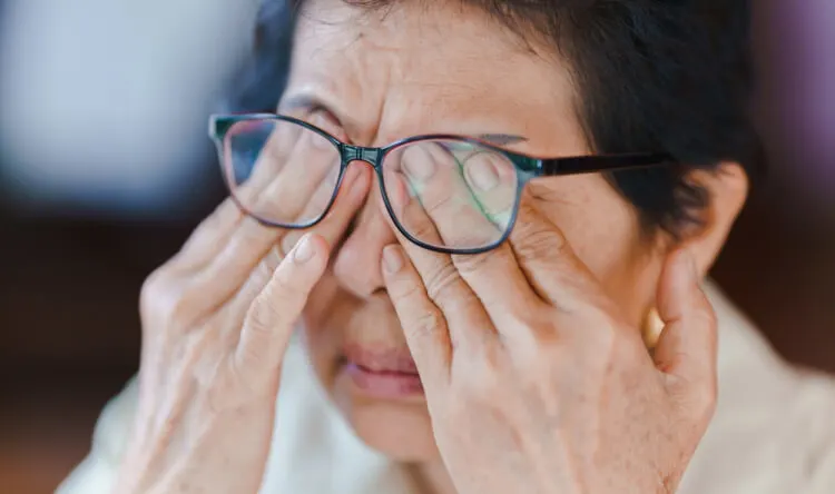 An elderly woman rubbing her eyes while wearing glasses, representing the need for supplements for vision support.